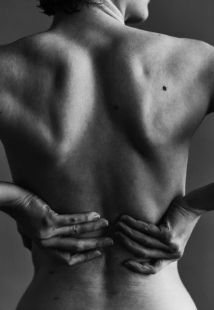Close-up black and white photo of a woman's back, hands resting on her lower back, showcasing skin texture and muscle definition against a plain backdrop.
