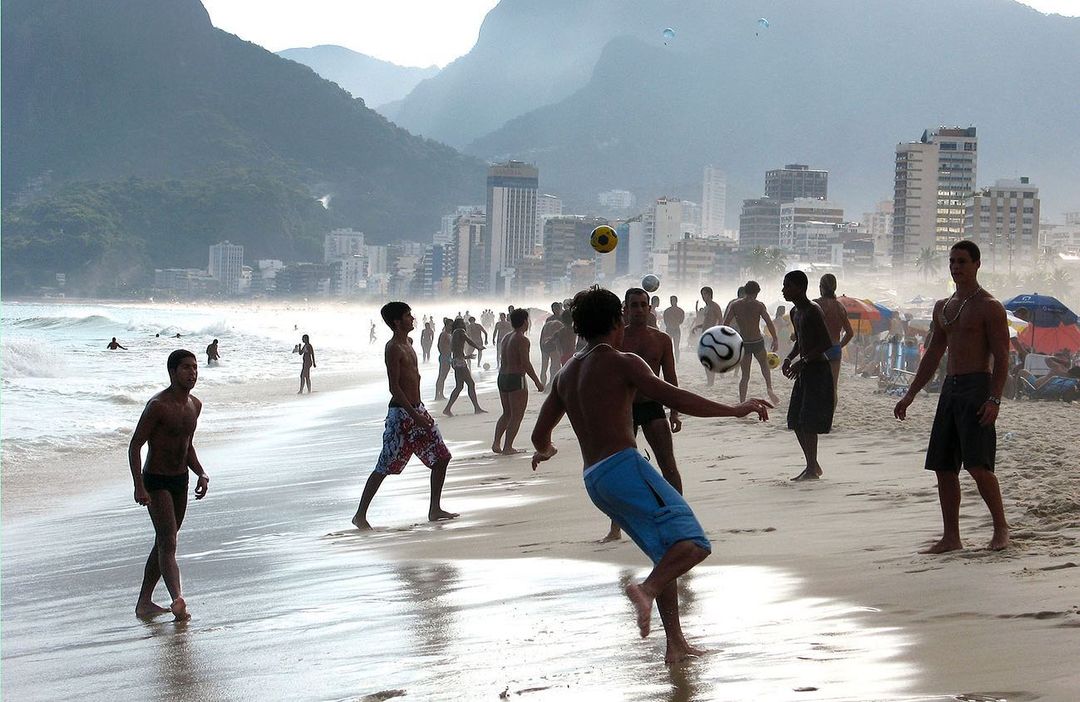 People playing soccer on a sandy beach with a city skyline and mountains in the background on a sunny day in an outdoor setting.
