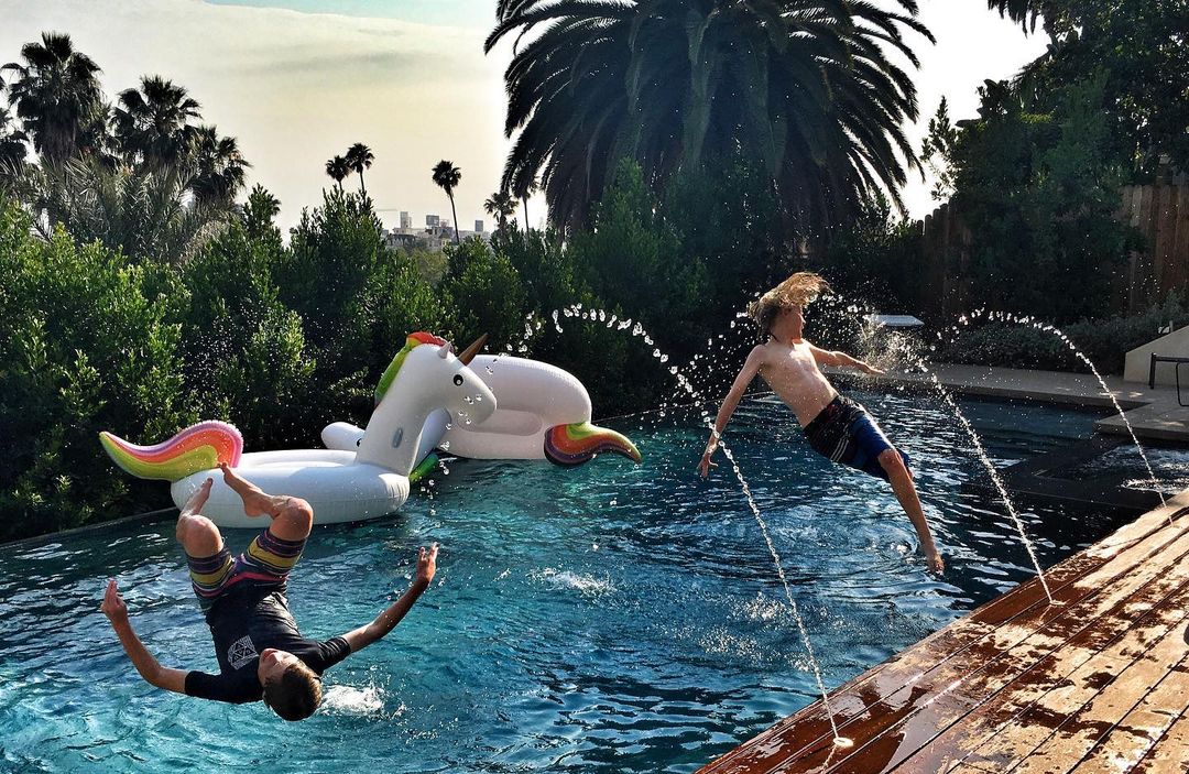 Two boys jumping into a pool near inflatable unicorn floats, surrounded by lush greenery and palm trees on a sunny day.