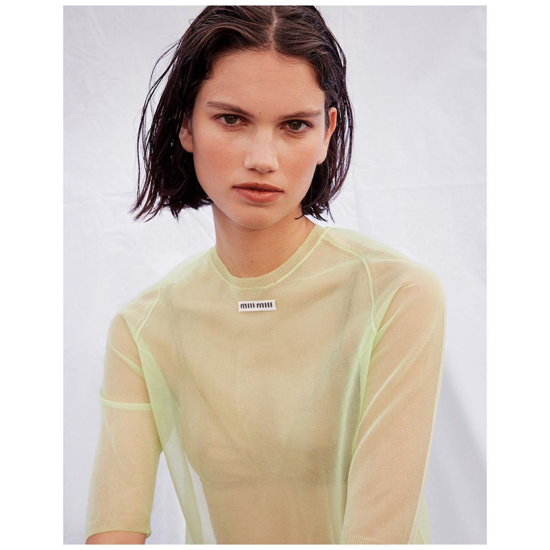 Fashion portrait of a brunette adult female wearing a neon green sheer long-sleeve top against a white background in a studio setting.