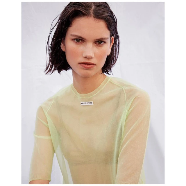 Fashion portrait of a brunette adult female wearing a neon green sheer long-sleeve top against a white background in a studio setting.