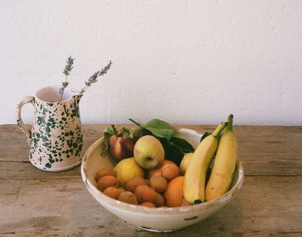 Still life photography featuring a fruit bowl filled with apricots, lemons, bananas and peaches, along with a jug with flowers.