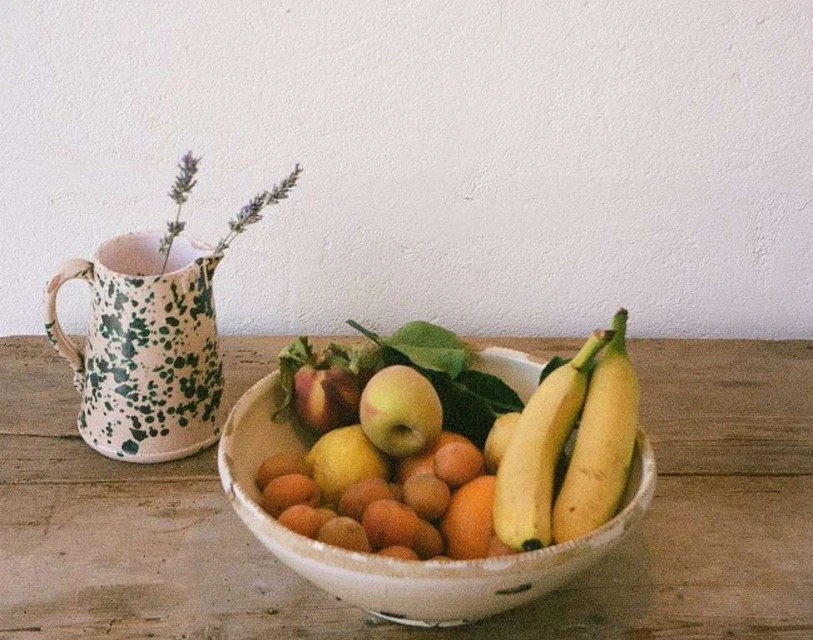 Still life photography featuring a fruit bowl filled with apricots, lemons, bananas and peaches, along with a jug with flowers.