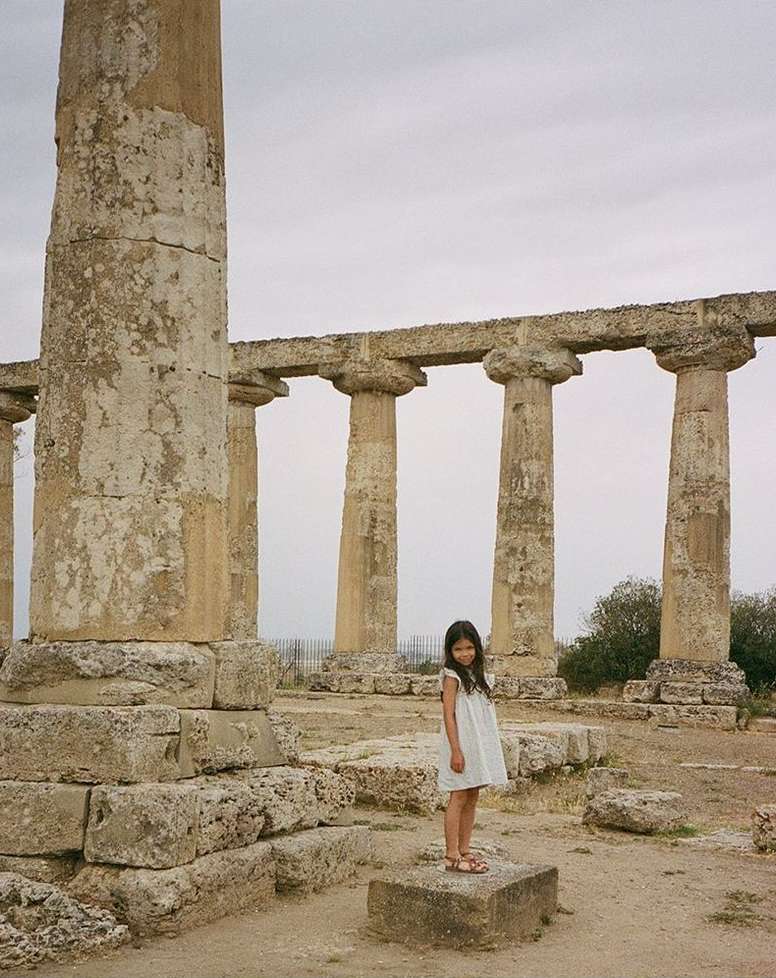 A young girl in a white dress stands on a stone block amidst the ruins of an ancient temple with massive columns under a cloudy sky.