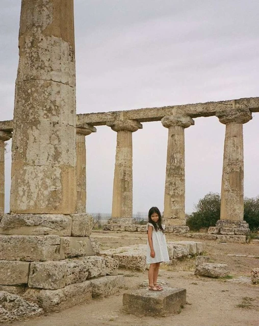 A young girl in a white dress stands on a stone block amidst the ruins of an ancient temple with massive columns under a cloudy sky.