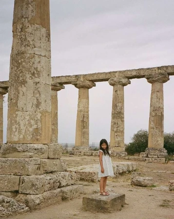 A young girl in a white dress stands on a stone block amidst the ruins of an ancient temple with massive columns under a cloudy sky.
