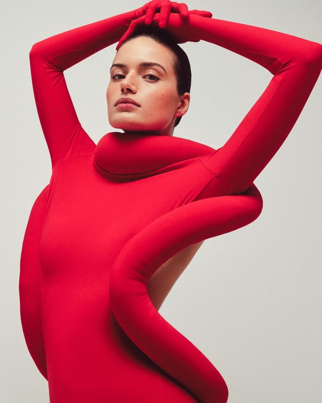 Woman in a red bodysuit poses with her arms raised above her head against a pale backdrop in a studio fashion shot.
