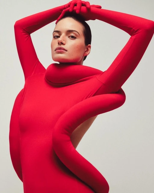 Woman in a red bodysuit poses with her arms raised above her head against a pale backdrop in a studio fashion shot.