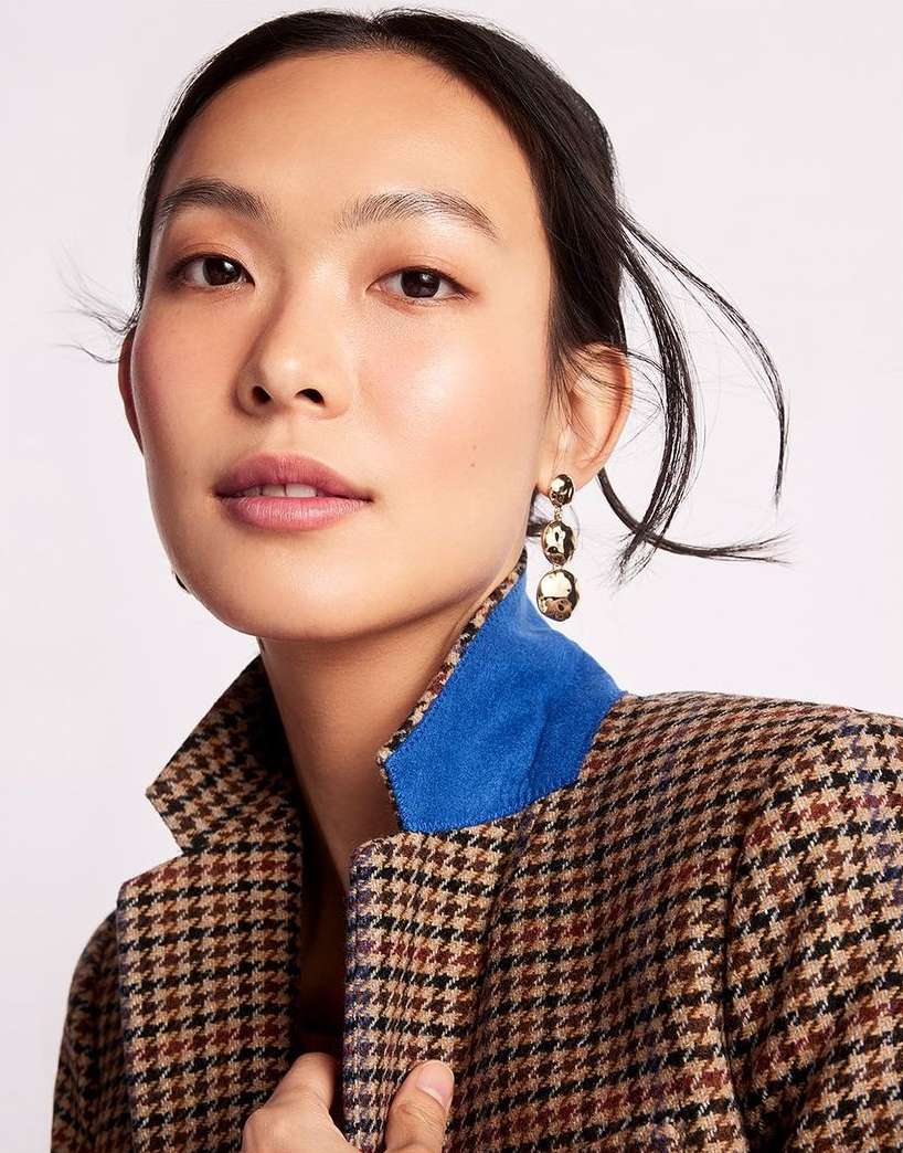 Portrait of a young Asian woman wearing a brown houndstooth jacket and gold triple-sphere earrings against a white backdrop.
