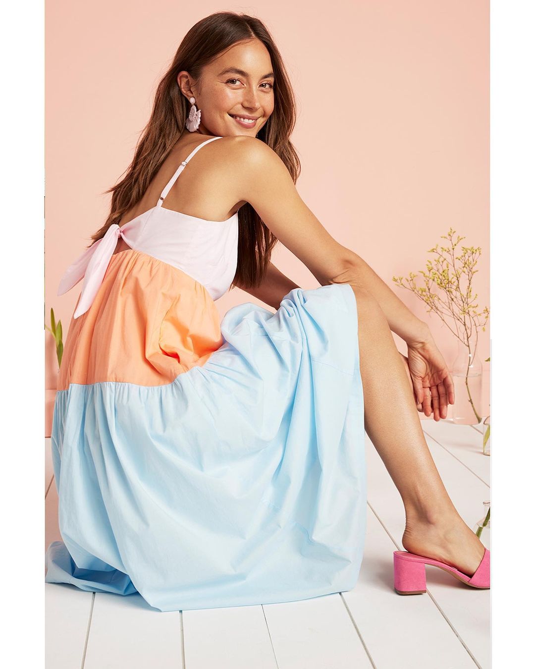 Attractive woman wearing a summery, color-blocked maxi dress in a studio setting, seated on a white wood floor, smiling at the camera.