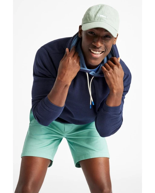 A dark-skinned male model in a blue hoodie, light-green shorts, and baseball cap poses in front of a white backdrop in a studio setting.