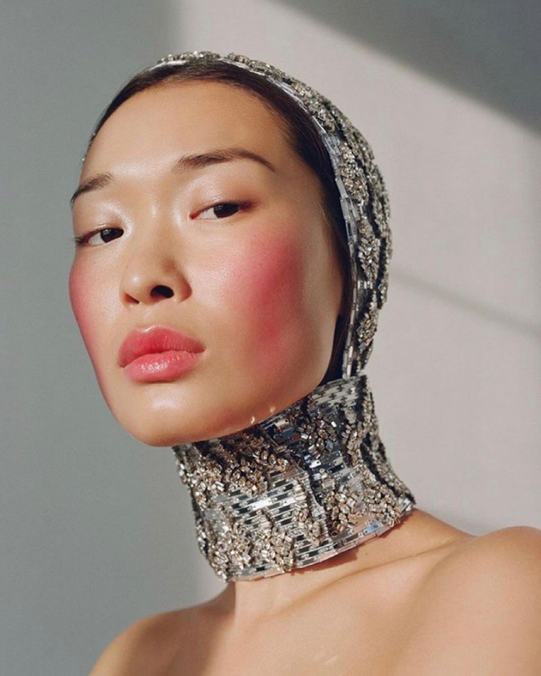 Close-up portrait of a woman with fair skin wearing a beaded head covering and choker, featuring bright pink blush and glossy lips.
