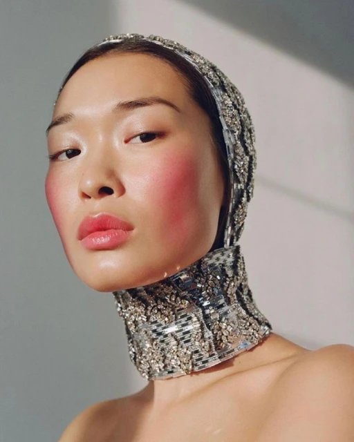Close-up portrait of a woman with fair skin wearing a beaded head covering and choker, featuring bright pink blush and glossy lips.