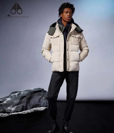 Black man wearing cream padded puffer jacket and black trousers standing in studio with a rock prop