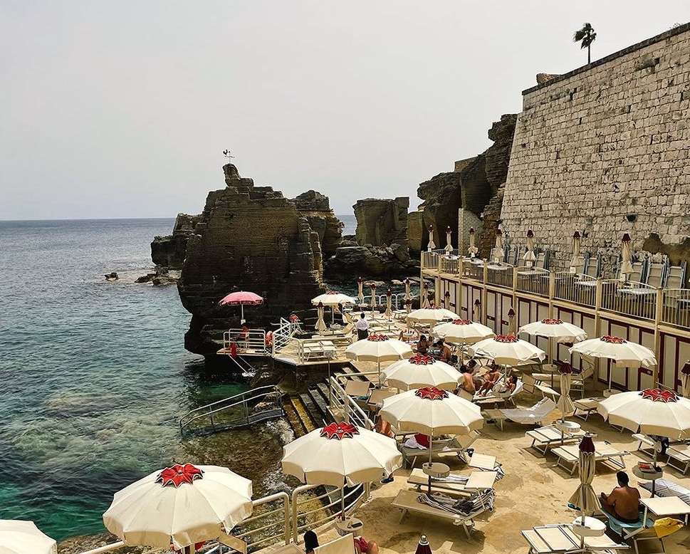 A sun-soaked beach scene with white umbrellas and lounge chairs lining the shore, set against a backdrop of rocky cliffs and turquoise water.