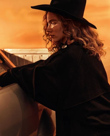 Woman in a black wide-brim hat and dark jacket posing beside a car against an orange sunset sky.
