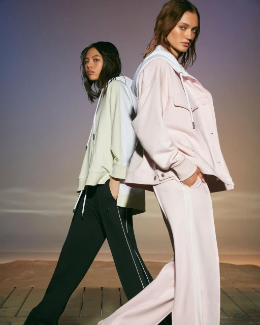 Two fashion models pose back to back wearing matching athletic jackets and pants in a photo studio with a desert-themed background.