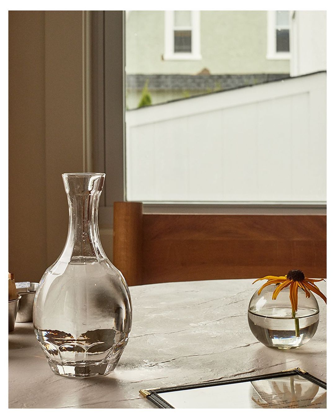 Still life of clear glass decanter with water, flower in glass vase, menu on marble-like table with building exterior in background.