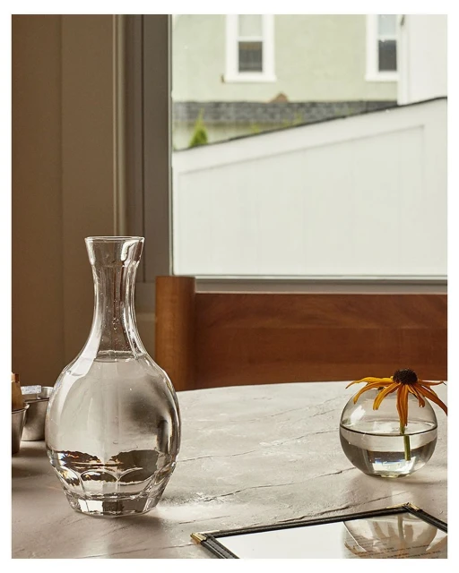 Still life of clear glass decanter with water, flower in glass vase, menu on marble-like table with building exterior in background.