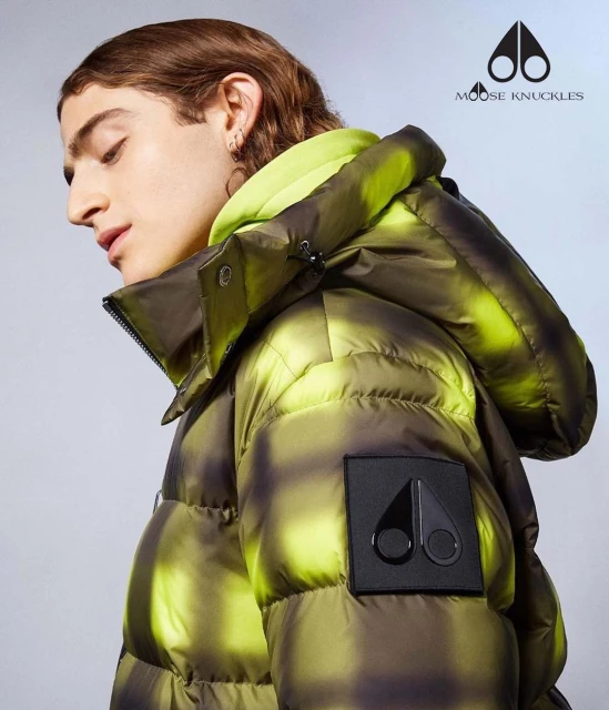 Adult male wearing a plaid puffer jacket by Moose Knuckles.  The jacket is green and black. He has brown hair and multiple earrings.