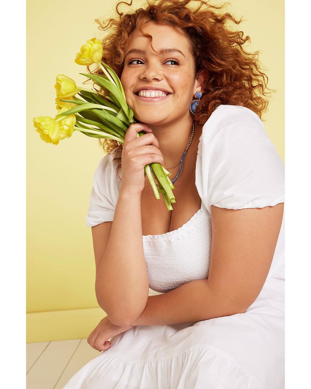 Smiling woman in a white dress sits while holding a bouquet of yellow tulips in front of a yellow background in a medium shot.