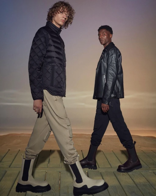 Two men pose in a studio wearing stylish boots, trousers, and jackets against a gradient background with an outdoor setting.