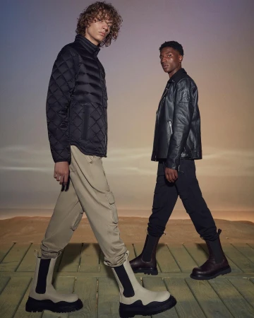 Two men pose in a studio wearing stylish boots, trousers, and jackets against a gradient background with an outdoor setting.