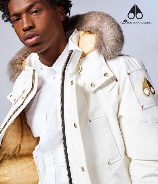 Close-up of a Black male model wearing a white fur-trimmed parka with gold hardware and a white collared shirt, showcasing Moose Knuckles logo.