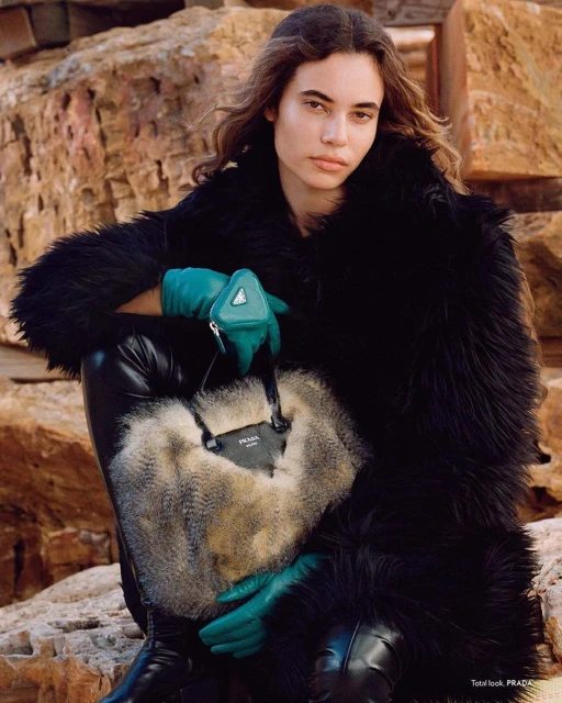 Fashion shot of a model wearing a black fur coat and leather pants, holding a fur and leather Prada purse, posing near rocks.