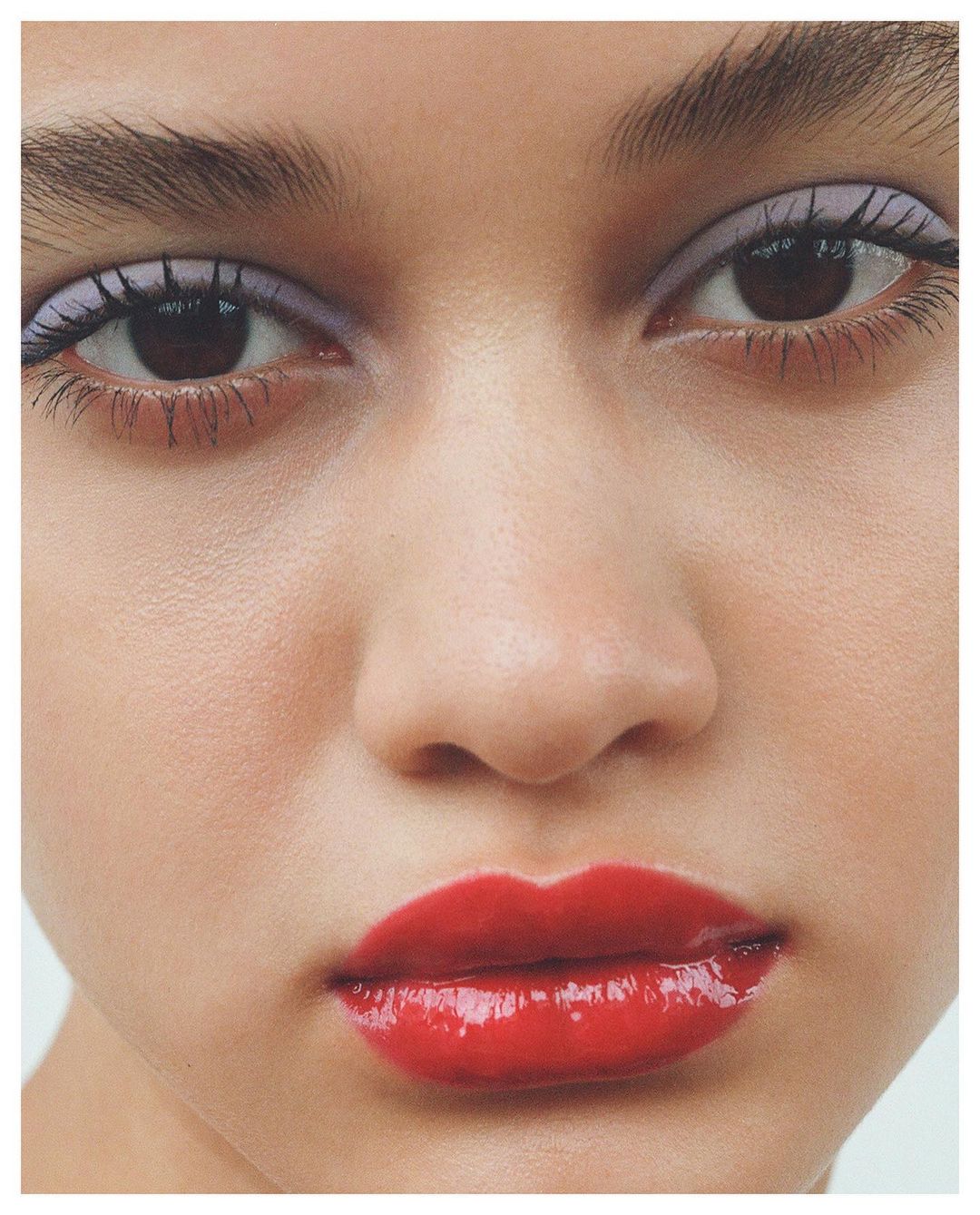 Close-up portrait of a woman with red lipstick and light purple eyeshadow, showcasing beauty and makeup in a clean, bright setting.