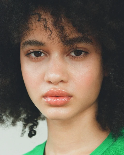 Close-up portrait of a young woman with curly dark hair, fair skin, and glossy lips wearing a green shirt against a neutral background.