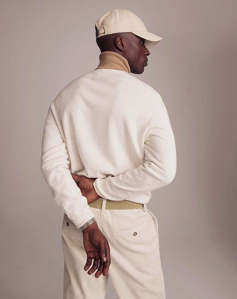 Man wearing a cream cap and white sweater with his hands behind his back against a neutral studio backdrop