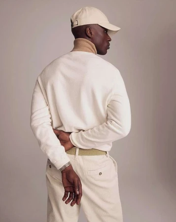 Man wearing a cream cap and white sweater with his hands behind his back against a neutral studio backdrop