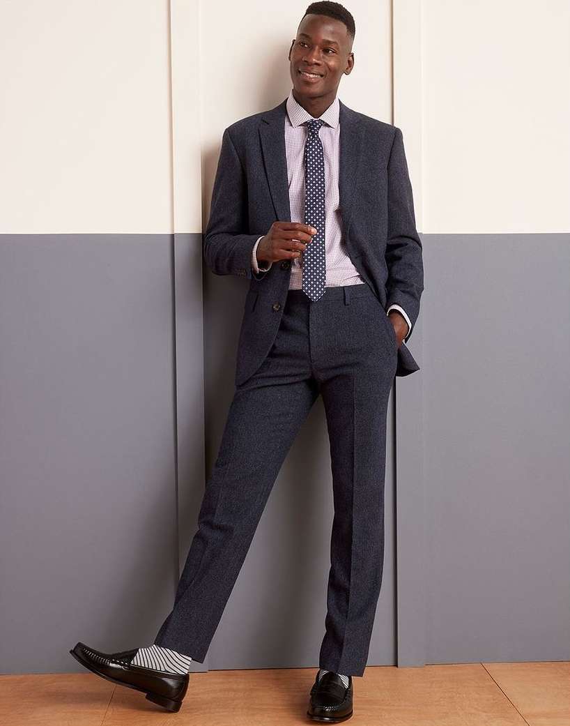 A young adult Black man in a suit poses against a wall, looking upward and to the side, wearing a tie and striped socks with shiny black shoes.