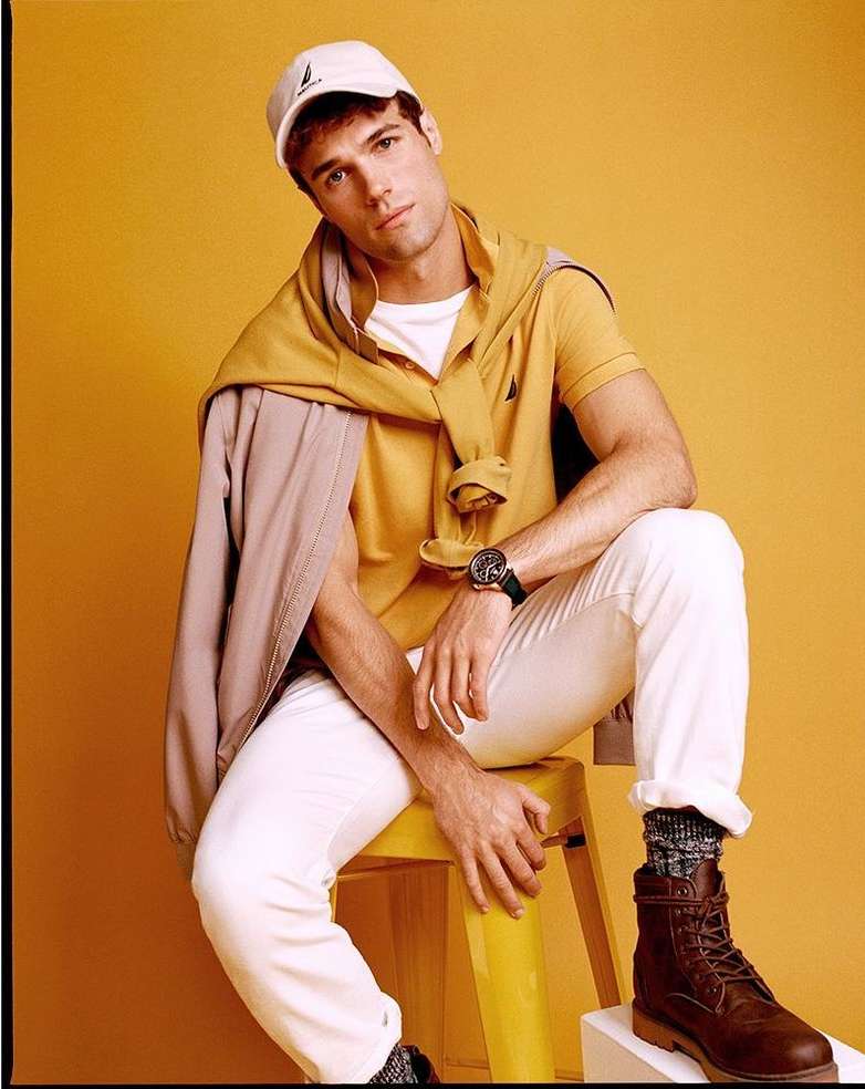 Young man sits on a yellow stool wearing white jeans, a yellow polo shirt, and brown boots against a yellow background.