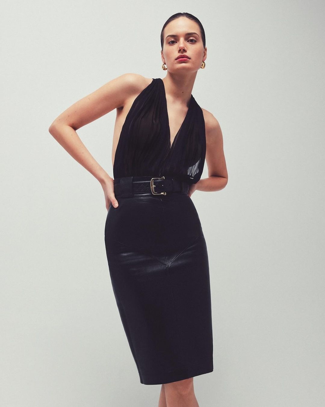 A woman poses in a black halter top and pencil skirt against a plain background, emphasizing fashion and minimalistic studio photography.