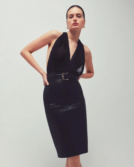 A woman poses in a black halter top and pencil skirt against a plain background, emphasizing fashion and minimalistic studio photography.