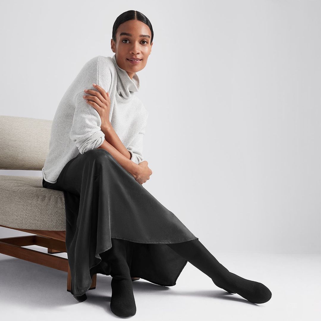 Adult woman sits on chair in studio, wearing a grey cowl-neck sweater, a black skirt, and black boots against a white background.