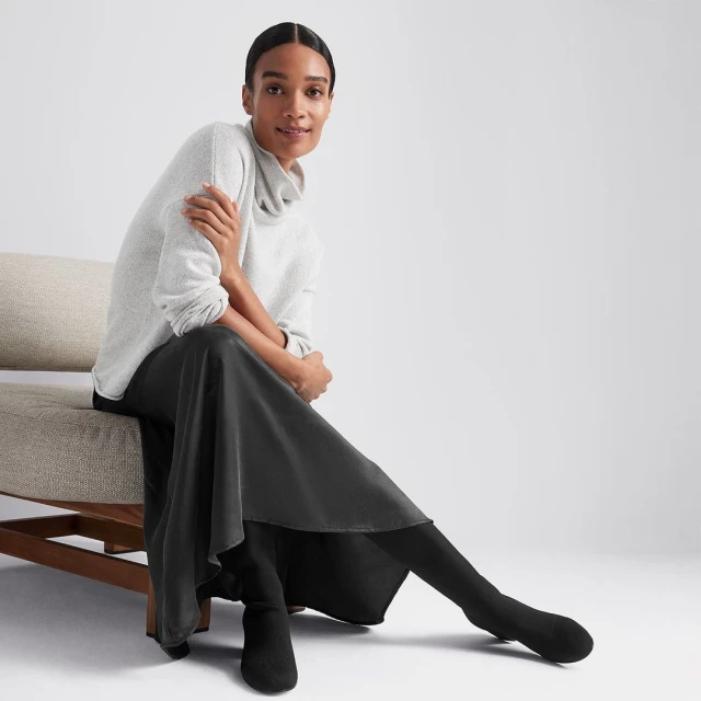 Adult woman sits on chair in studio, wearing a grey cowl-neck sweater, a black skirt, and black boots against a white background.