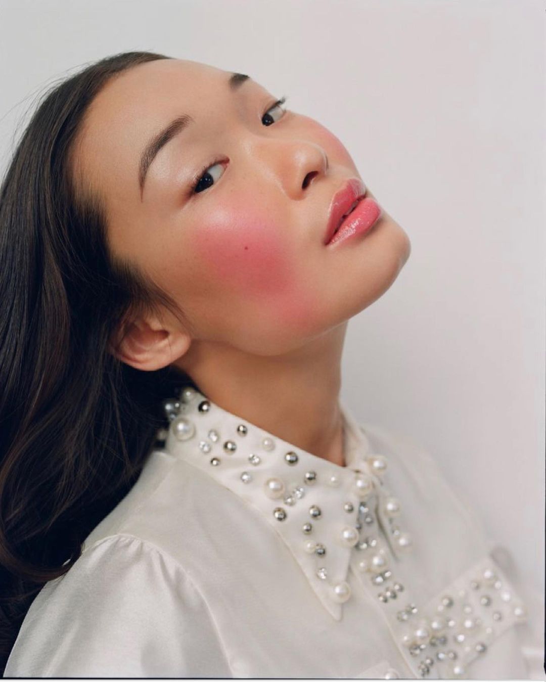 Portrait of an Asian woman with fair skin and red blush, wearing a white pearl-beaded satin shirt, angled against a white backdrop.