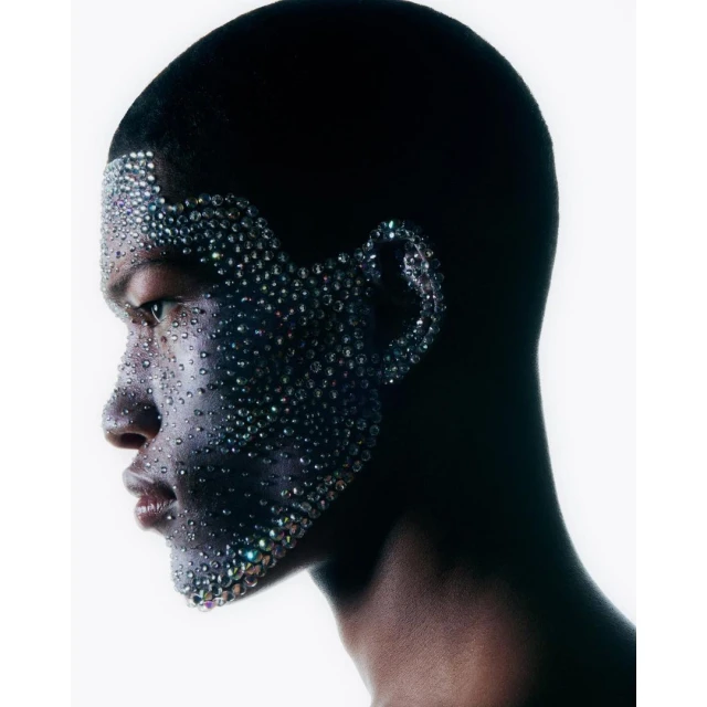 Dramatic profile portrait of a Black adult woman with crystal embellishments on her face against a bright, high-key background.