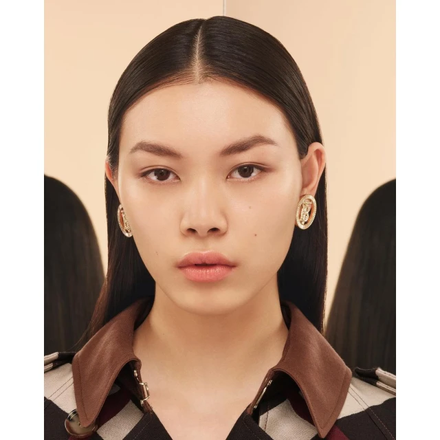 Close-up of an Asian woman with dark hair wearing gold oval logo earrings and a brown, red, and white patterned top in a studio.