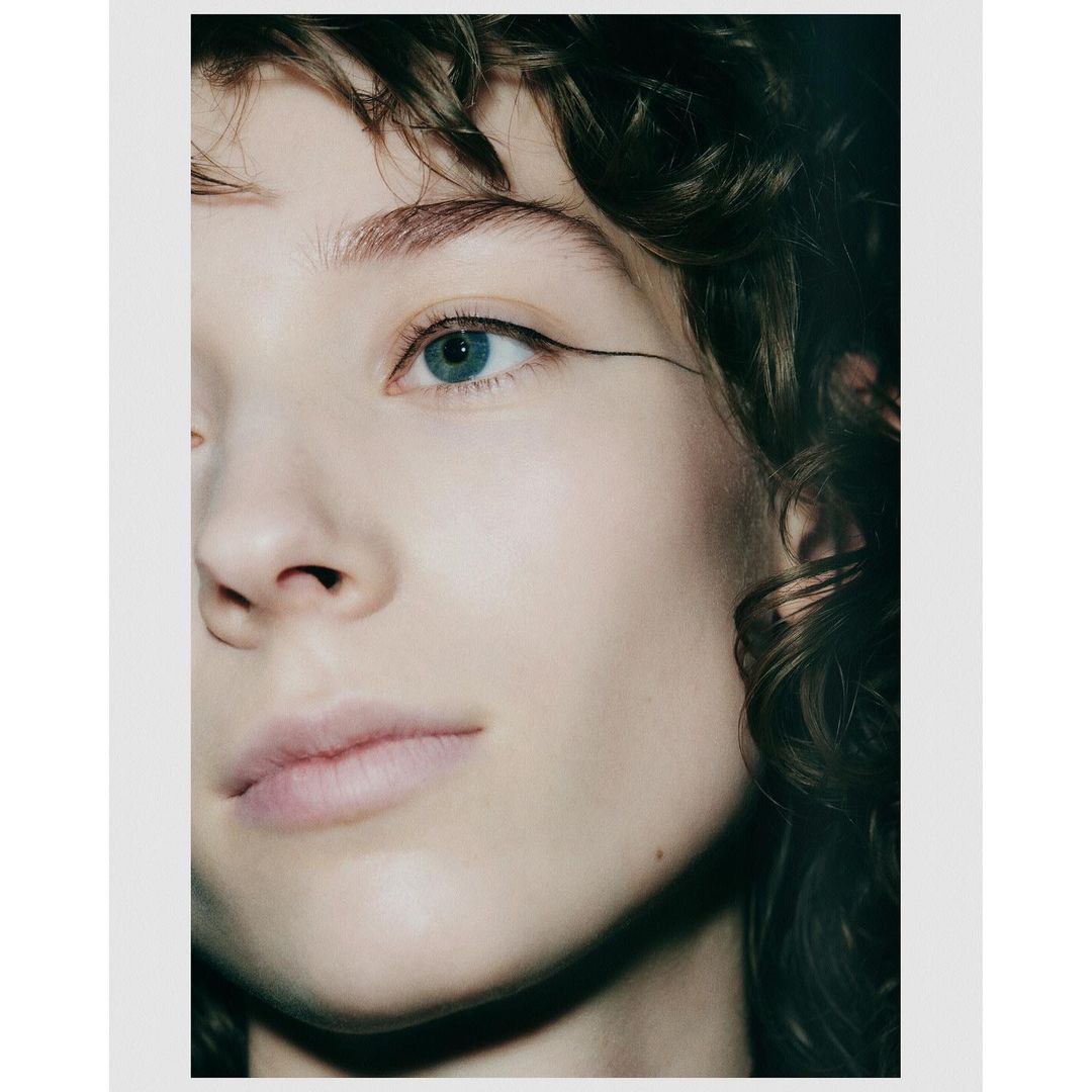 Close-up portrait of a young fair-skinned woman with an expressive gaze, dark curly hair, and winged eyeliner against a soft, neutral background.