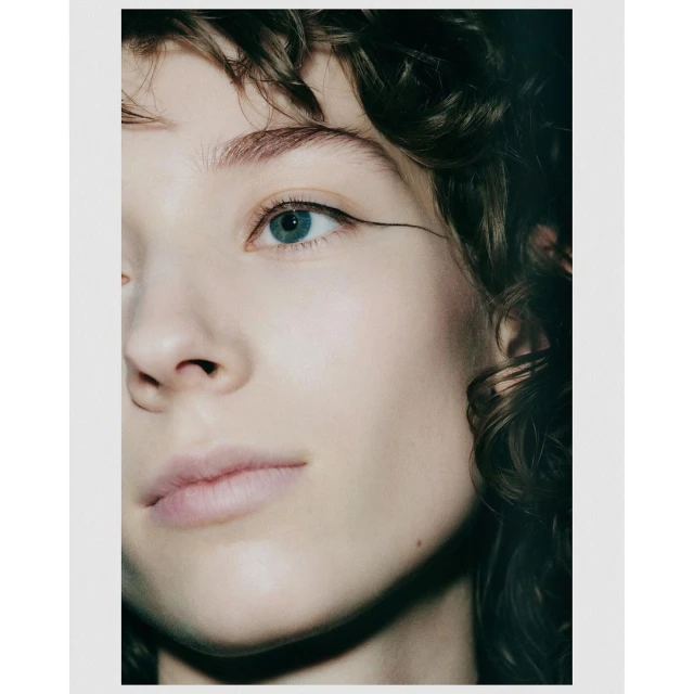 Close-up portrait of a young fair-skinned woman with an expressive gaze, dark curly hair, and winged eyeliner against a soft, neutral background.