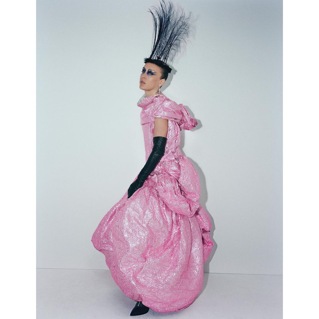 Adult female model in a pink shimmering couture gown with black gloves and feathered headpiece in a full studio shot on white.