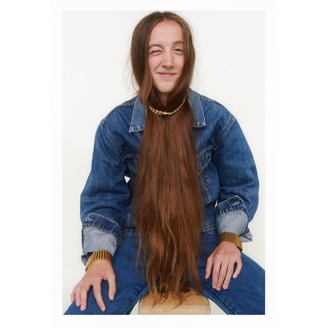 Portrait of a woman wearing a blue denim jacket with long brown hair covering her face and winking at the camera against a white backdrop.