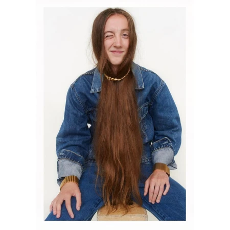 Portrait of a woman wearing a blue denim jacket with long brown hair covering her face and winking at the camera against a white backdrop.