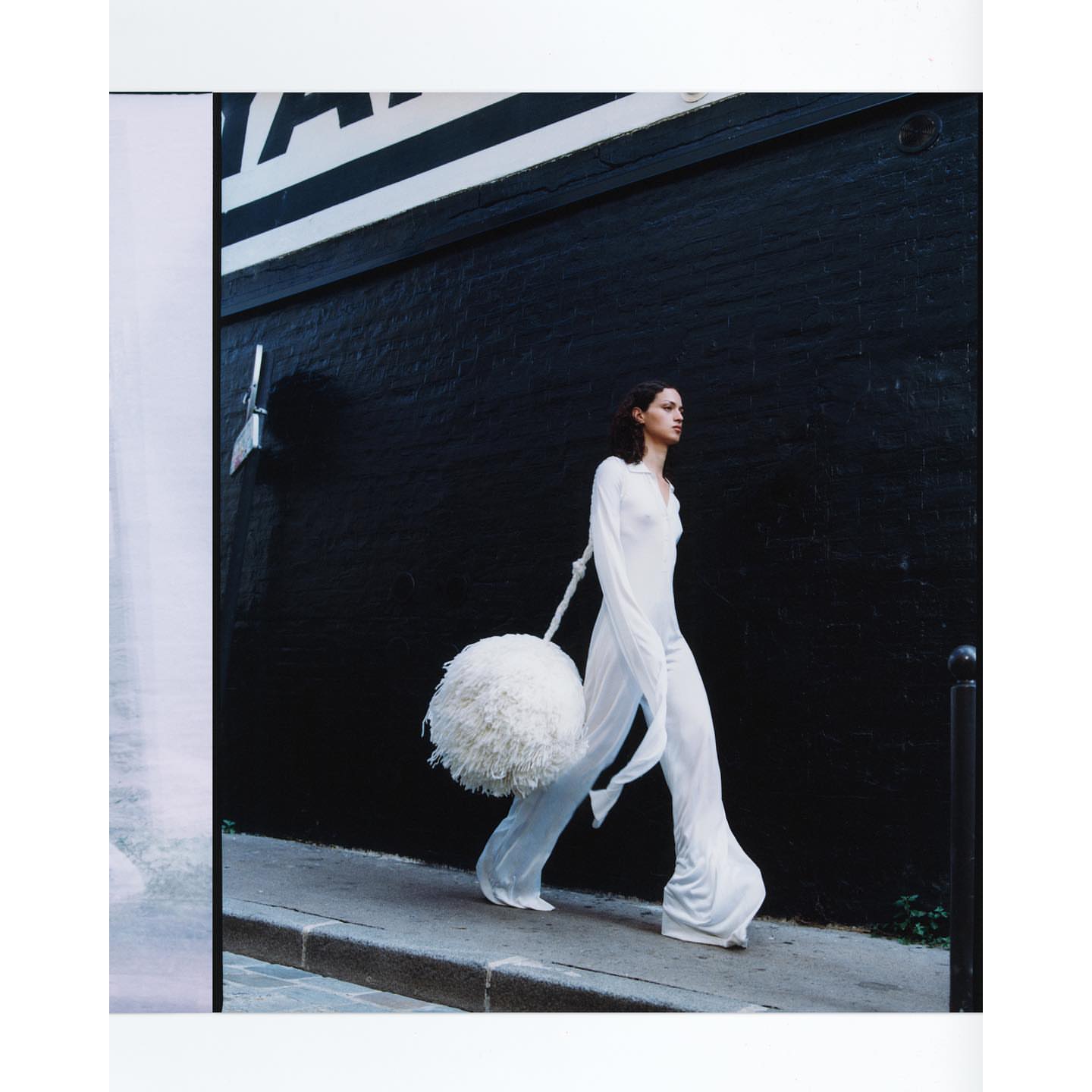 Fashion shot of a slender adult female wearing a long white dress and carrying a large white pompom while walking along a city street.