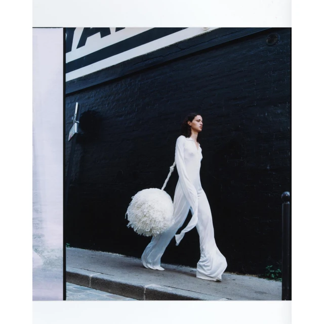 Fashion shot of a slender adult female wearing a long white dress and carrying a large white pompom while walking along a city street.