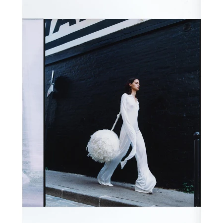 Fashion shot of a slender adult female wearing a long white dress and carrying a large white pompom while walking along a city street.
